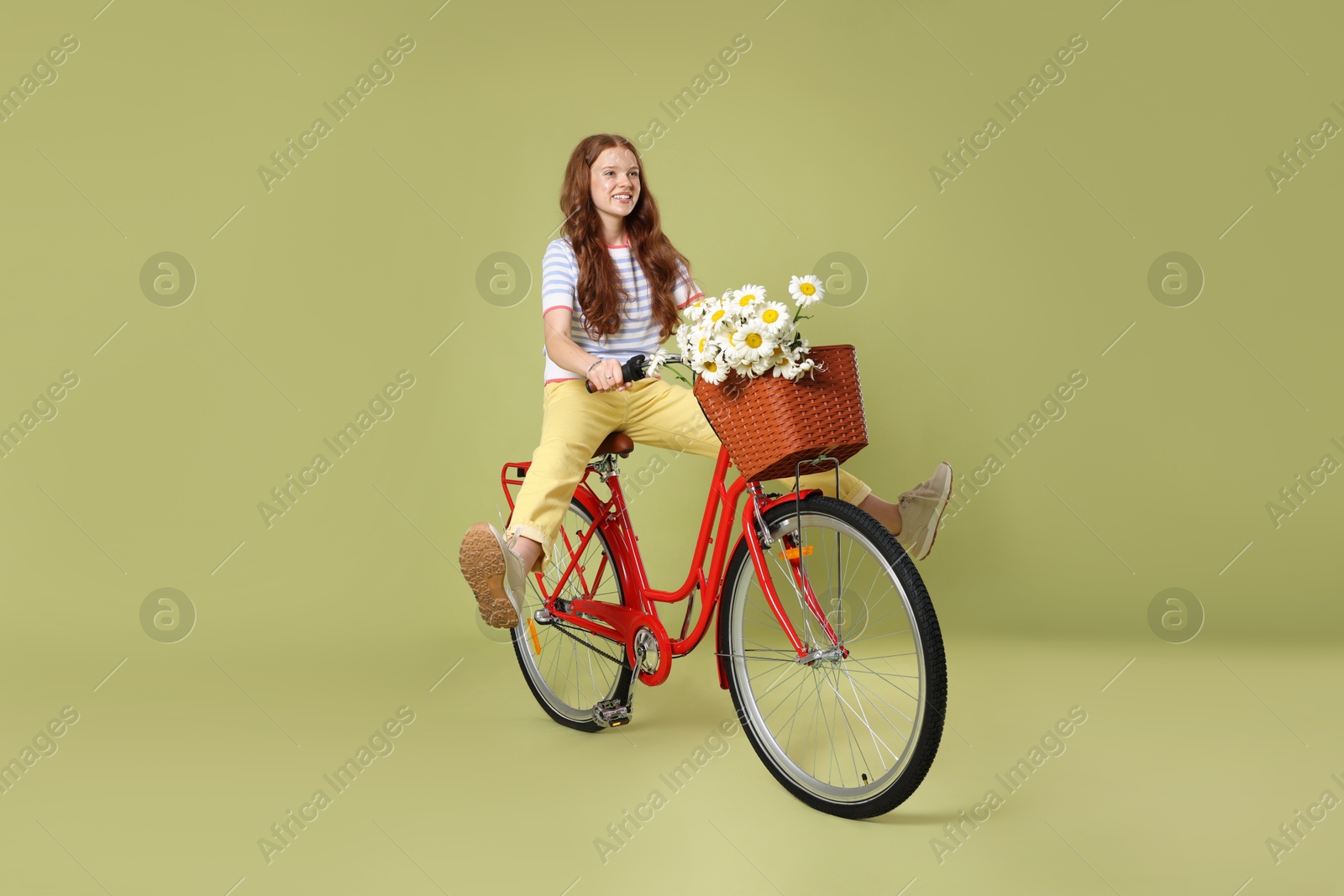 Smiling teenage girl riding bicycle with basket of flowers against green background Photo of Smiling teenage girl riding bicycle with basket of flowers against green background
