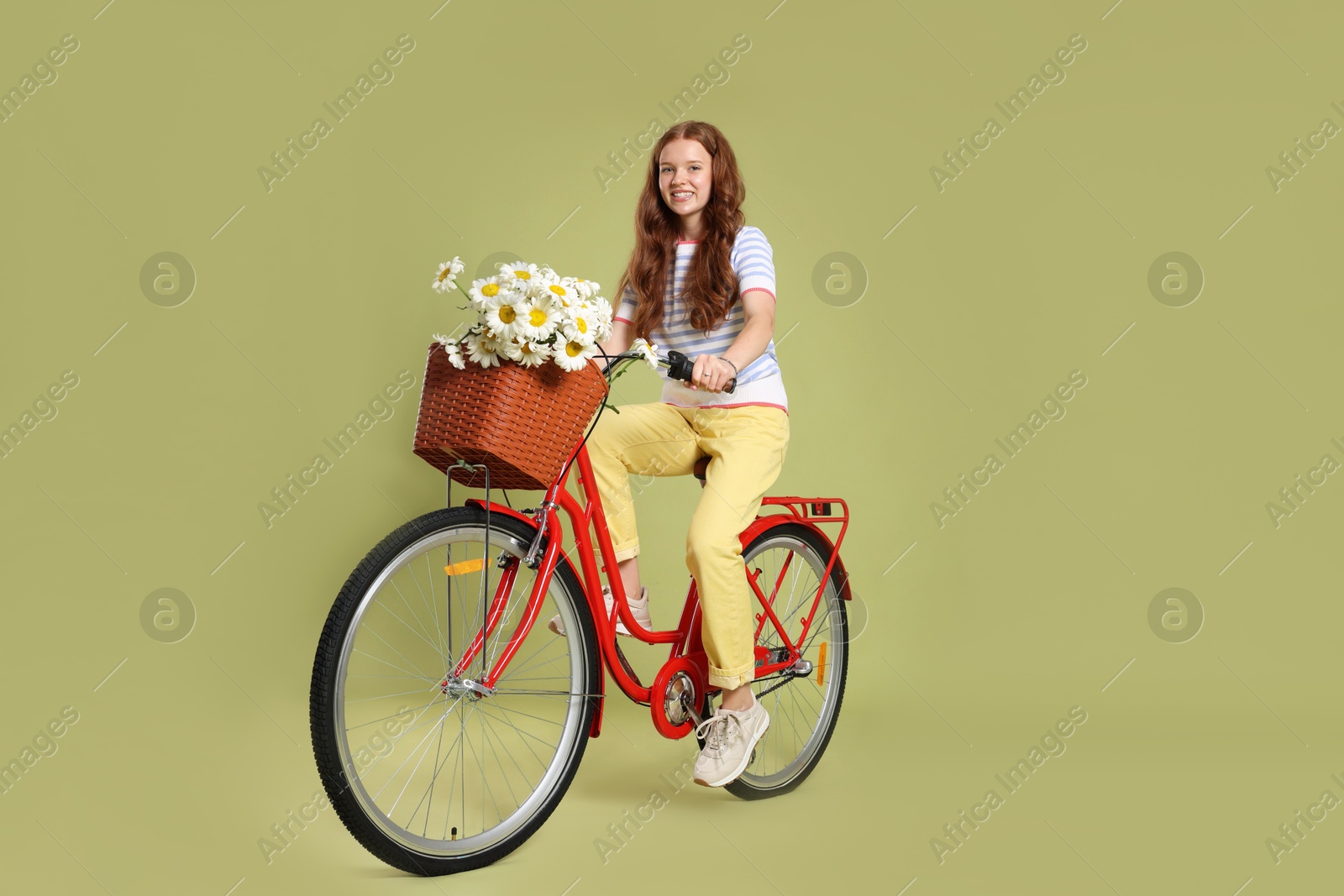 Smiling teenage girl riding bicycle with basket of flowers against green background Photo of Smiling teenage girl riding bicycle with basket of flowers against green background