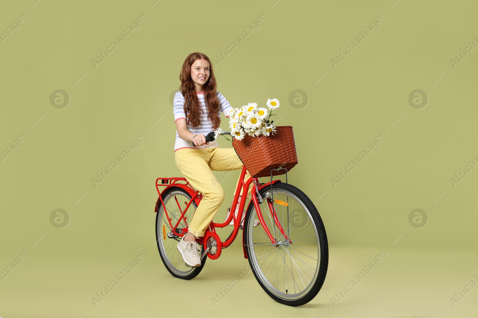 Smiling teenage girl riding bicycle with basket of flowers against green background Photo of Smiling teenage girl riding bicycle with basket of flowers against green background