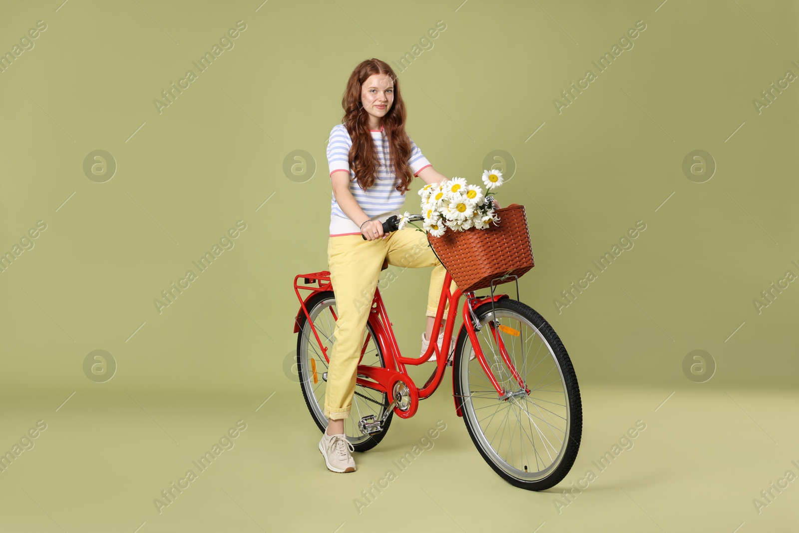Beautiful teenage girl riding bicycle with basket of flowers against green background Photo of Beautiful teenage girl riding bicycle with basket of flowers against green background