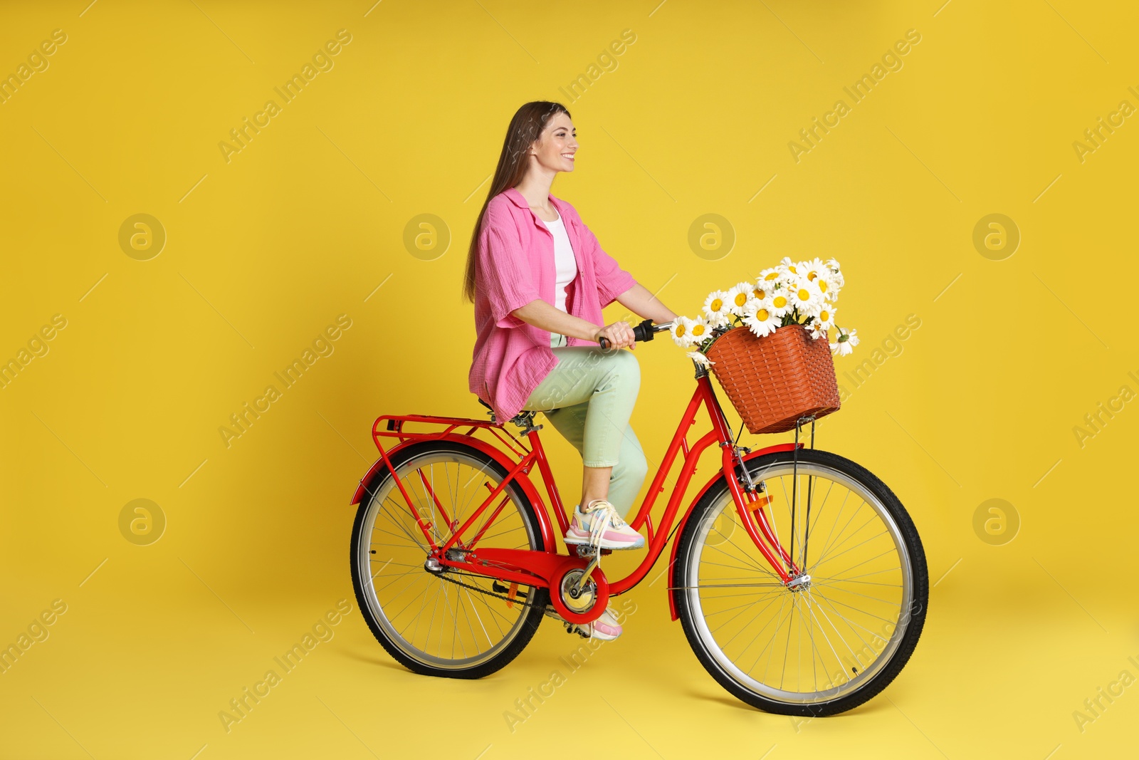 Photo of Smiling woman riding bicycle with basket of flowers against yellow background