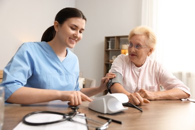 Photo of Nurse measuring patient's blood pressure at wooden table indoors. Home health care service