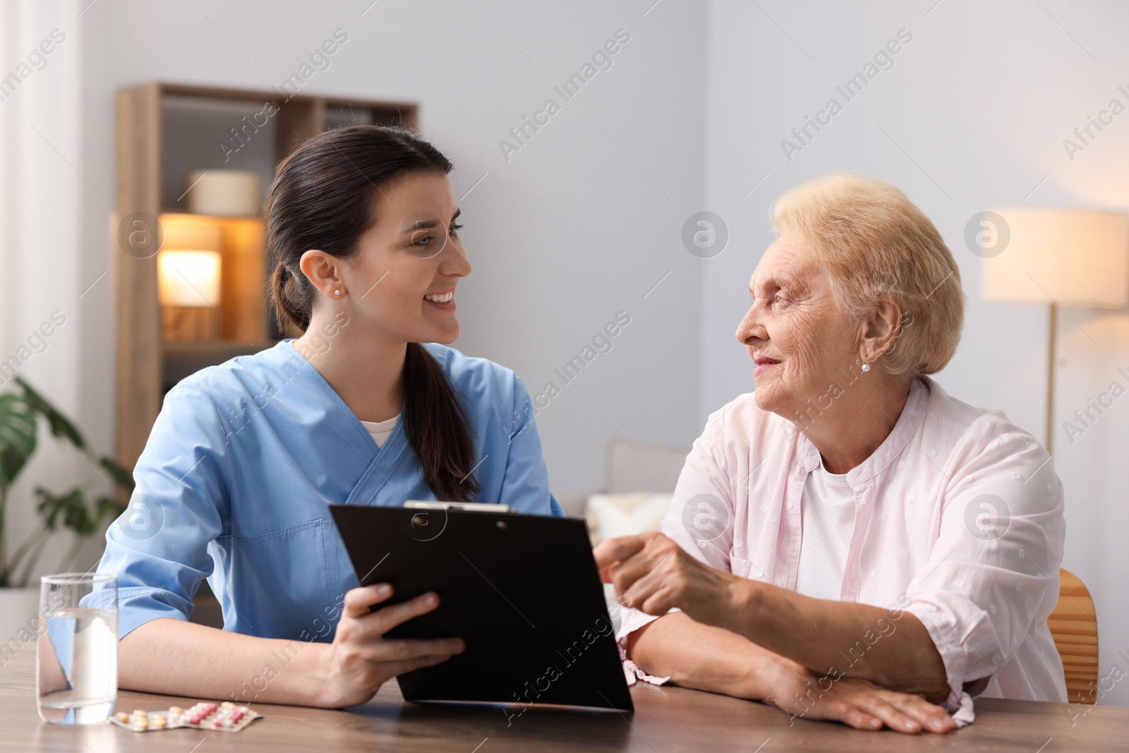 Nurse with clipboard examining senior woman at wooden table indoors. Home health care service Photo of Nurse with clipboard examining senior woman at wooden table indoors. Home health care service