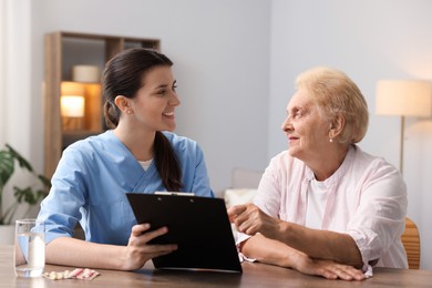 Photo of Nurse with clipboard examining senior woman at wooden table indoors. Home health care service
