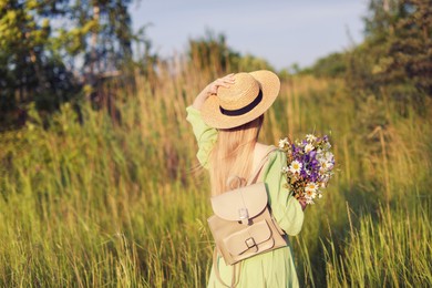 Photo of Woman with beautiful wildflowers, hat and backpack in meadow on sunny day, back view