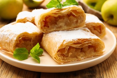 Photo of Pieces of tasty apple strudel with powdered sugar, mint and fruits on wooden table, closeup