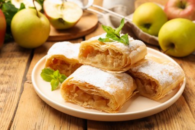 Photo of Pieces of tasty apple strudel with powdered sugar, mint and fruits on wooden table, closeup