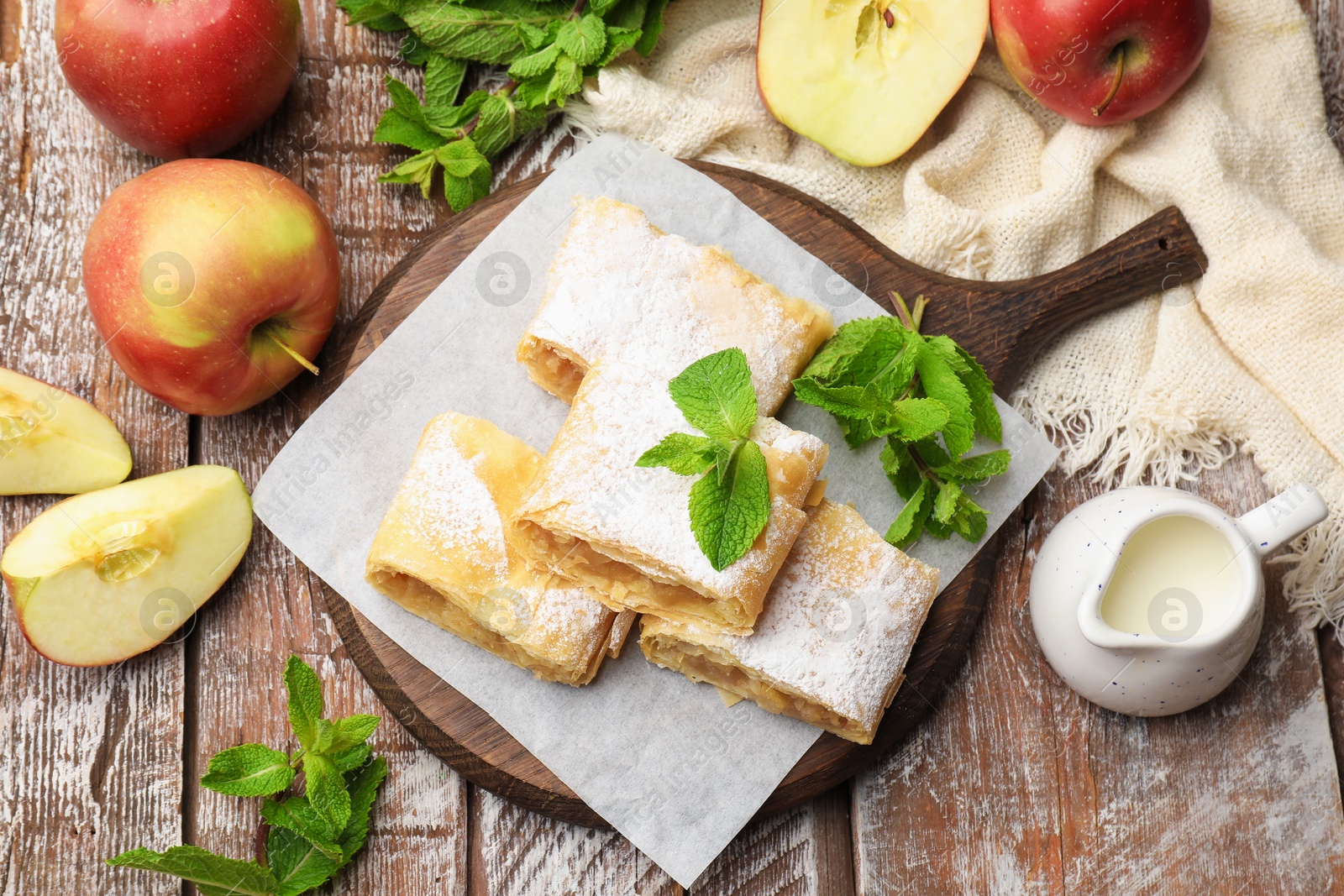 Pieces of tasty apple strudel with powdered sugar, mint and fruits on wooden table, flat lay Photo of Pieces of tasty apple strudel with powdered sugar, mint and fruits on wooden table, flat lay