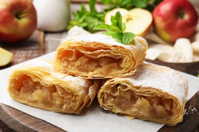 Photo of Pieces of tasty apple strudel with powdered sugar, mint and fruits on wooden table, closeup