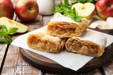 Photo of Pieces of tasty apple strudel with powdered sugar, mint and fruits on wooden table, closeup