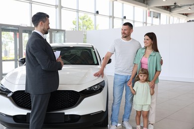 Photo of Salesman with clipboard showing new car to happy family in salon