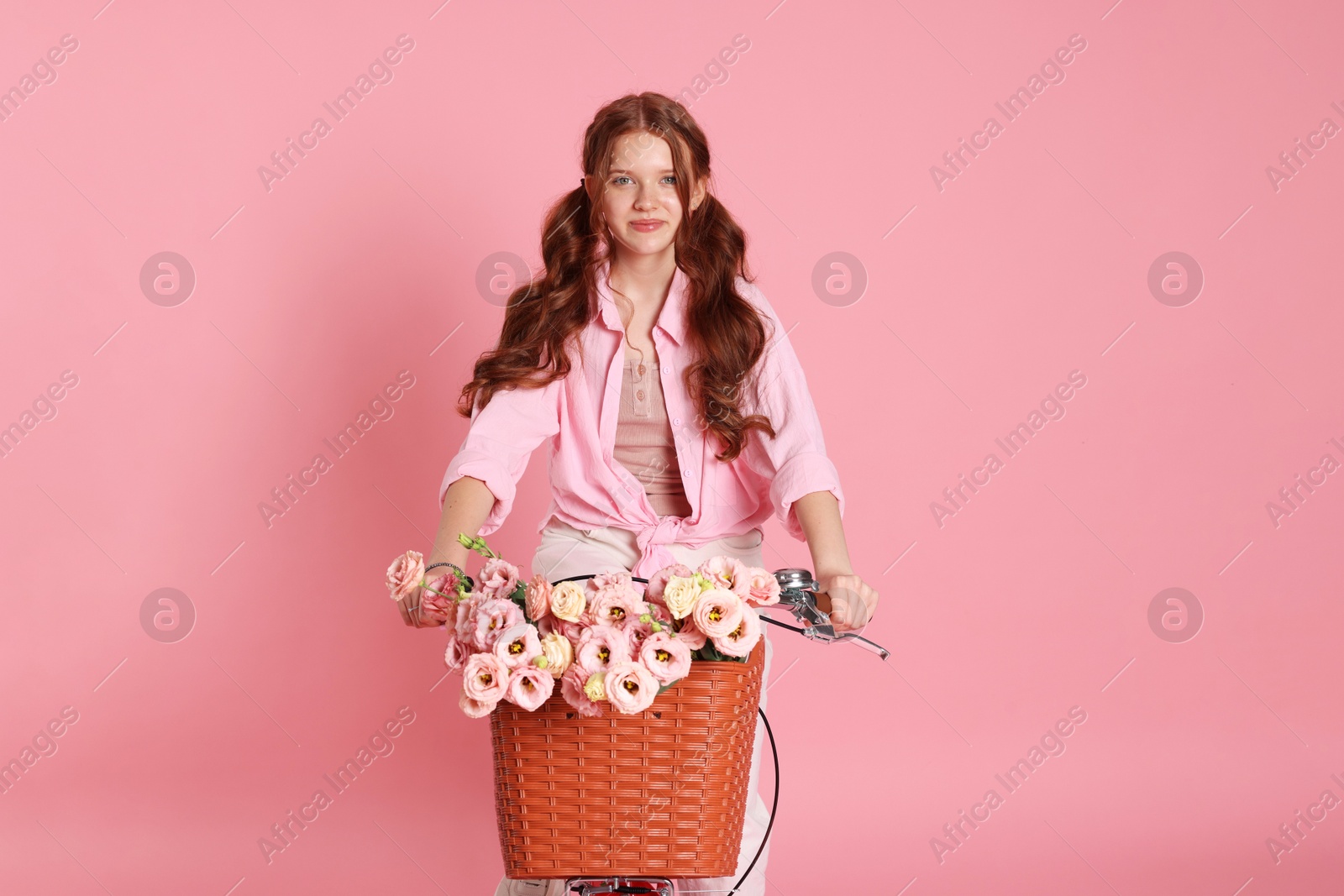 Photo of Beautiful teenage girl riding bicycle with basket of flowers on pink background
