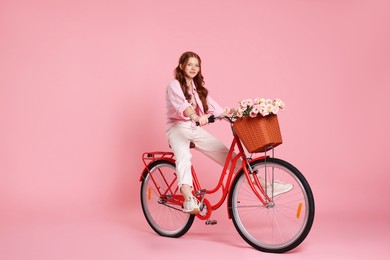 Photo of Beautiful teenage girl riding bicycle with basket of flowers on pink background. Space for text