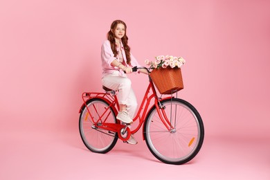 Photo of Beautiful teenage girl riding bicycle with basket of flowers on pink background