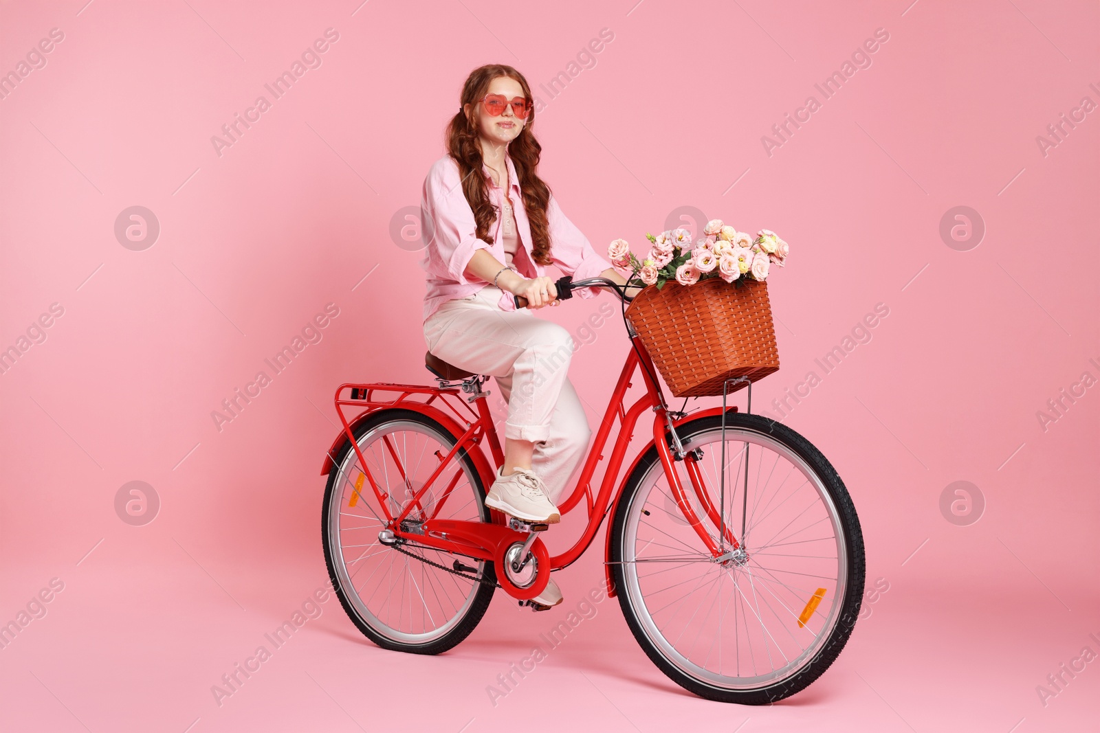 Beautiful teenage girl in sunglasses riding bicycle with basket of flowers on pink background Photo of Beautiful teenage girl in sunglasses riding bicycle with basket of flowers on pink background