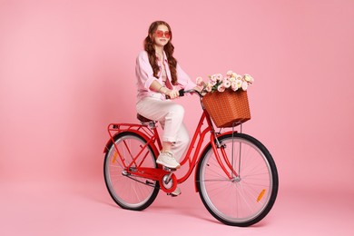 Photo of Beautiful teenage girl in sunglasses riding bicycle with basket of flowers on pink background