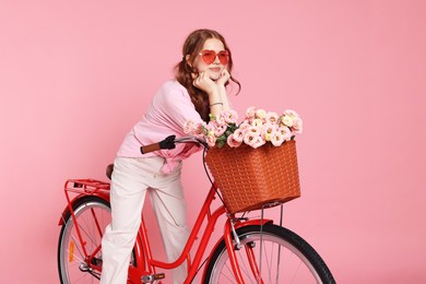Photo of Beautiful teenage girl in sunglasses posing on bicycle with basket of flowers against pink background
