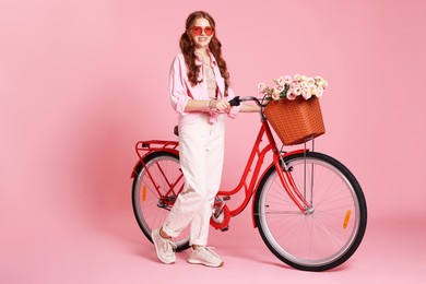 Photo of Smiling teenage girl in sunglasses and bicycle with basket of flowers against pink background. Space for text