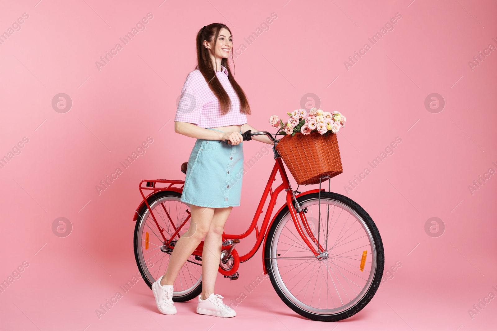 Smiling woman and bicycle with basket of flowers against pink background Photo of Smiling woman and bicycle with basket of flowers against pink background