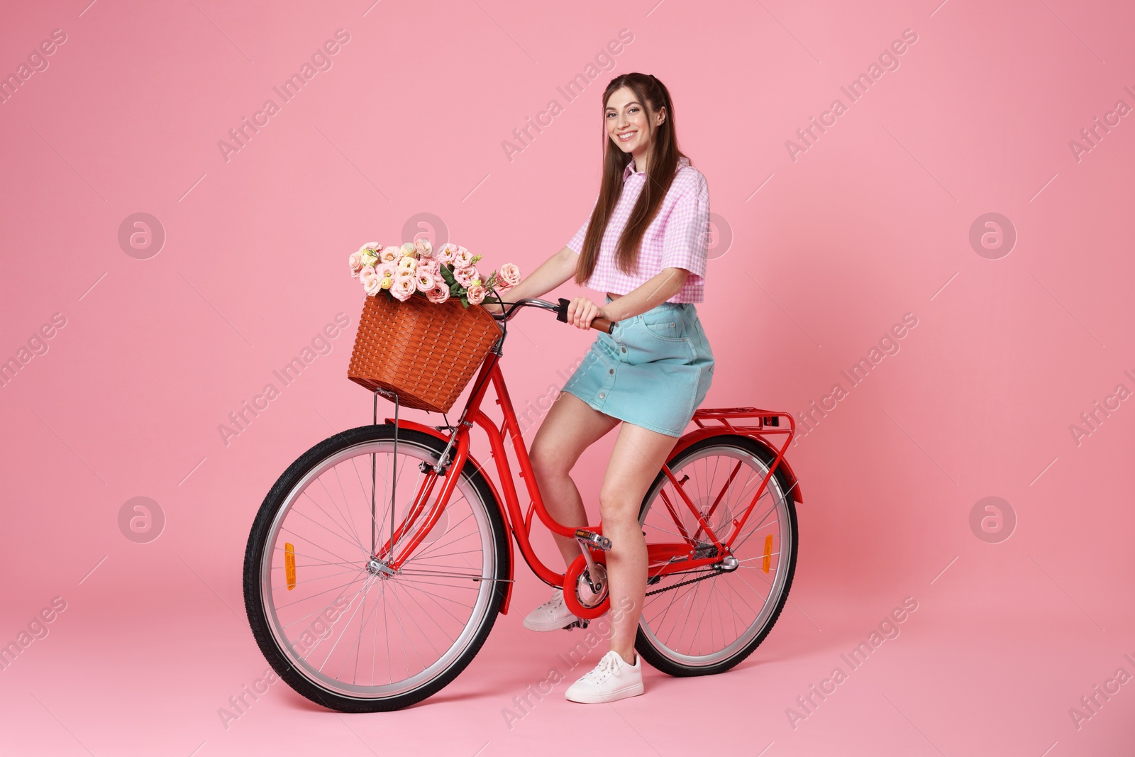 Photo of Smiling woman posing on bicycle with basket of flowers against pink background
