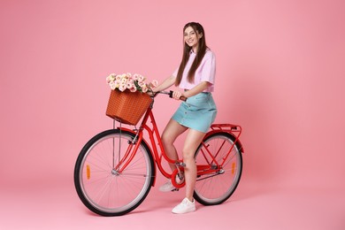 Photo of Smiling woman posing on bicycle with basket of flowers against pink background