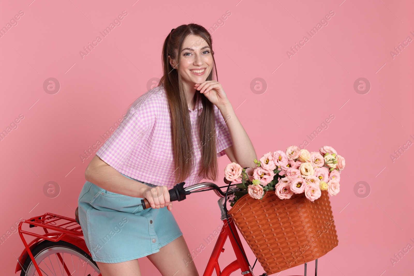 Smiling woman posing on bicycle with basket of flowers against pink background Photo of Smiling woman posing on bicycle with basket of flowers against pink background