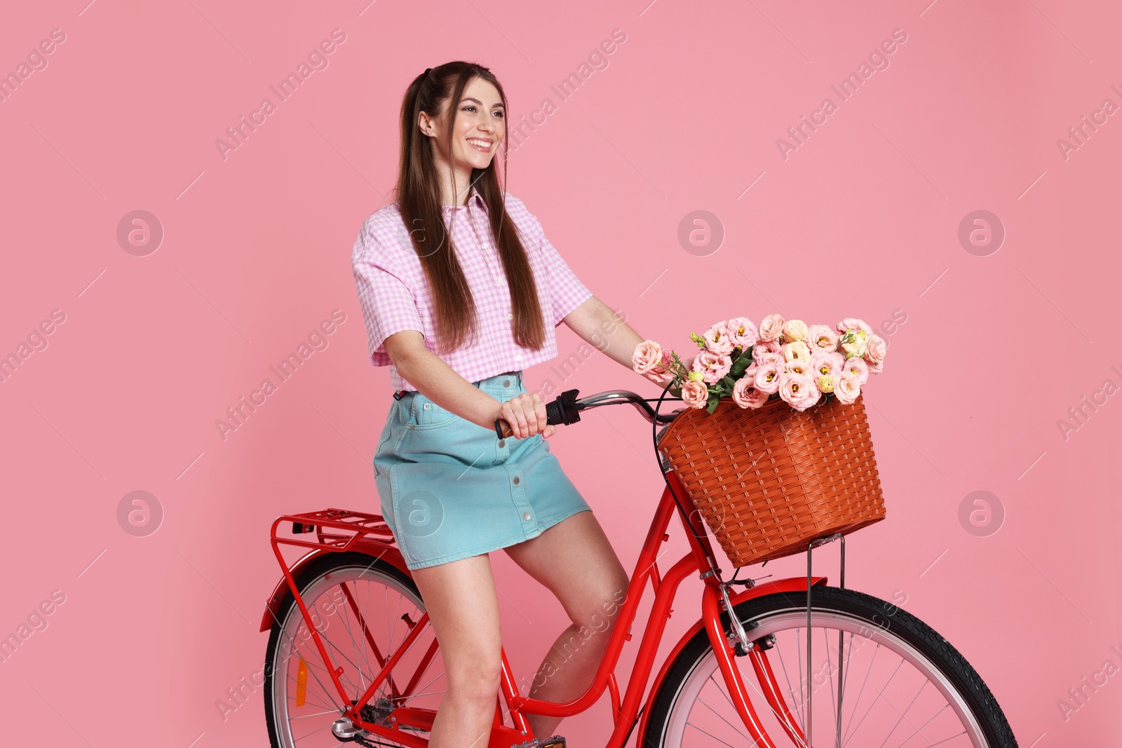 Smiling woman posing on bicycle with basket of flowers against pink background Photo of Smiling woman posing on bicycle with basket of flowers against pink background