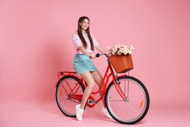 Photo of Smiling woman posing on bicycle with basket of flowers against pink background