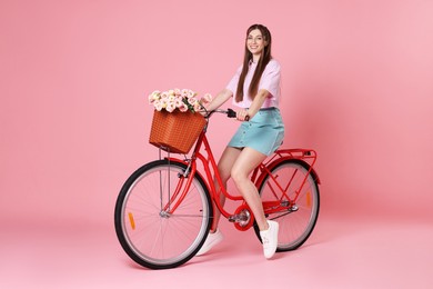 Photo of Smiling woman posing on bicycle with basket of flowers against pink background