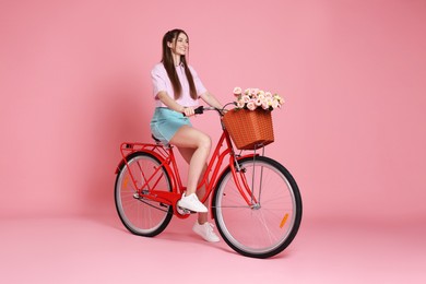 Photo of Smiling woman posing on bicycle with basket of flowers against pink background