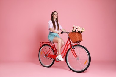 Photo of Smiling woman posing on bicycle with basket of flowers against pink background
