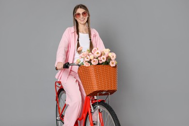 Photo of Smiling woman in sunglasses riding bicycle with basket of flowers against grey background