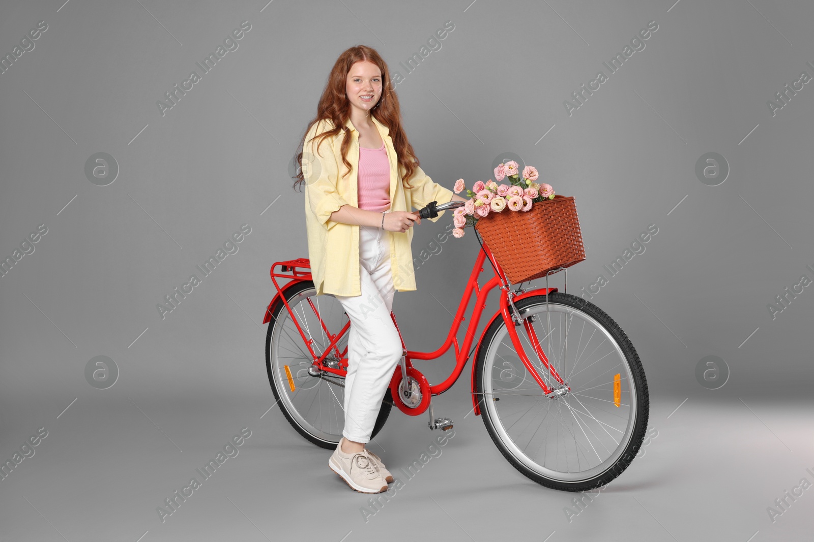 Smiling teenage girl and bicycle with basket of flowers against grey background Photo of Smiling teenage girl and bicycle with basket of flowers against grey background