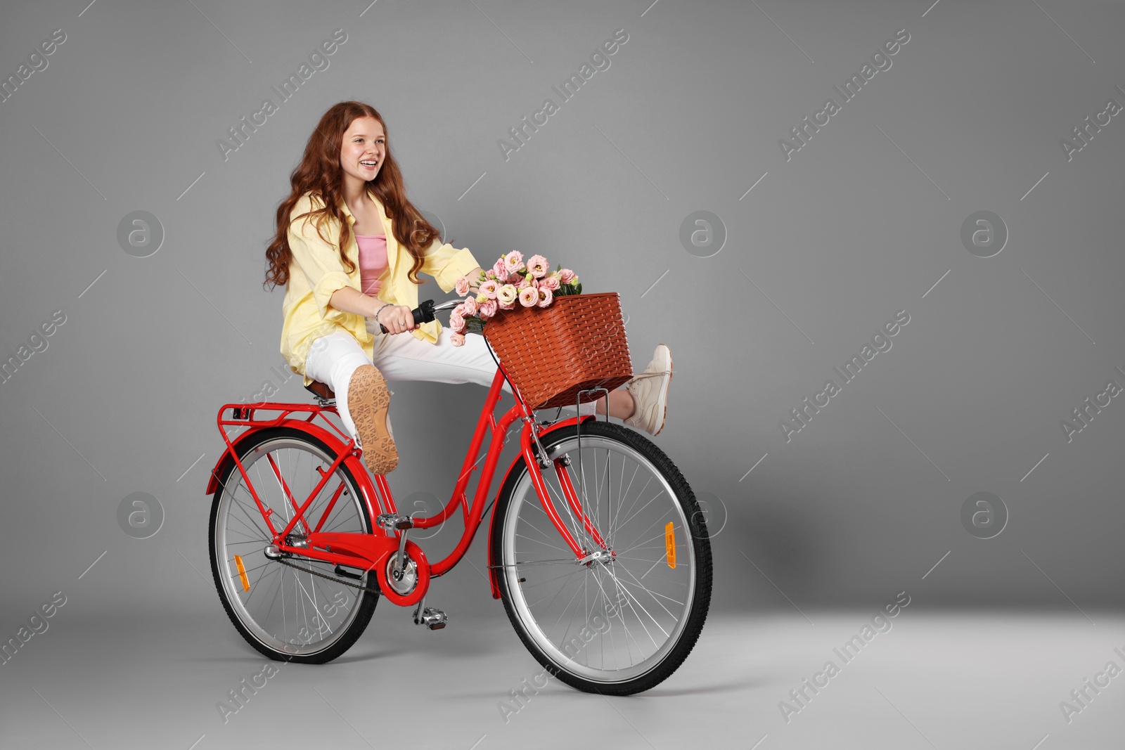Smiling teenage girl riding bicycle with basket of flowers against grey background Photo of Smiling teenage girl riding bicycle with basket of flowers against grey background