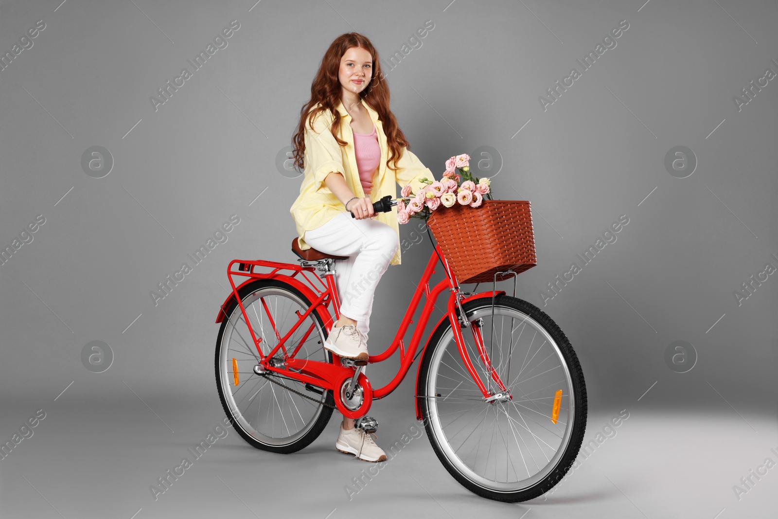 Beautiful teenage girl riding bicycle with basket of flowers against grey background Photo of Beautiful teenage girl riding bicycle with basket of flowers against grey background