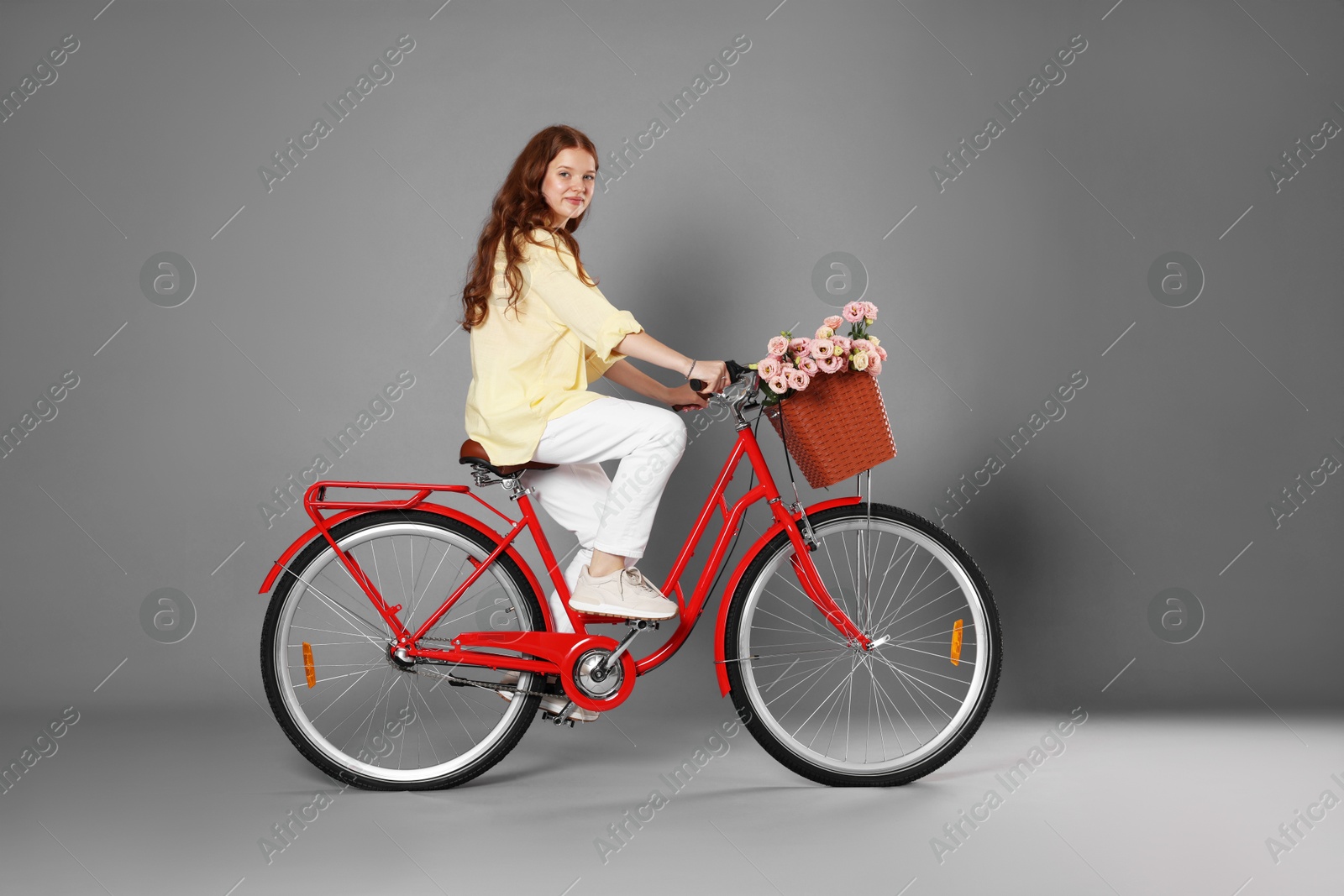 Beautiful teenage girl riding bicycle with basket of flowers against grey background Photo of Beautiful teenage girl riding bicycle with basket of flowers against grey background