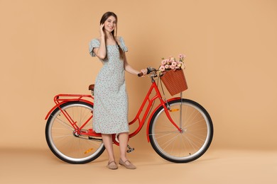 Photo of Smiling woman in dress and bicycle with basket of flowers on beige background