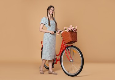 Photo of Smiling woman in dress and bicycle with basket of flowers on beige background
