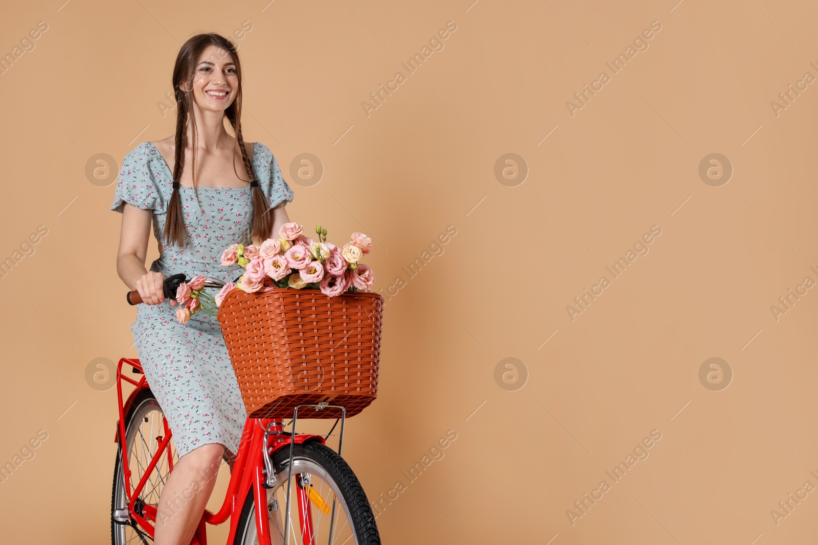 Smiling woman in dress riding bicycle with basket of flowers on beige background. Space for text Photo of Smiling woman in dress riding bicycle with basket of flowers on beige background. Space for text