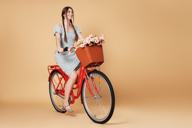 Photo of Smiling woman in dress riding bicycle with basket of flowers on beige background