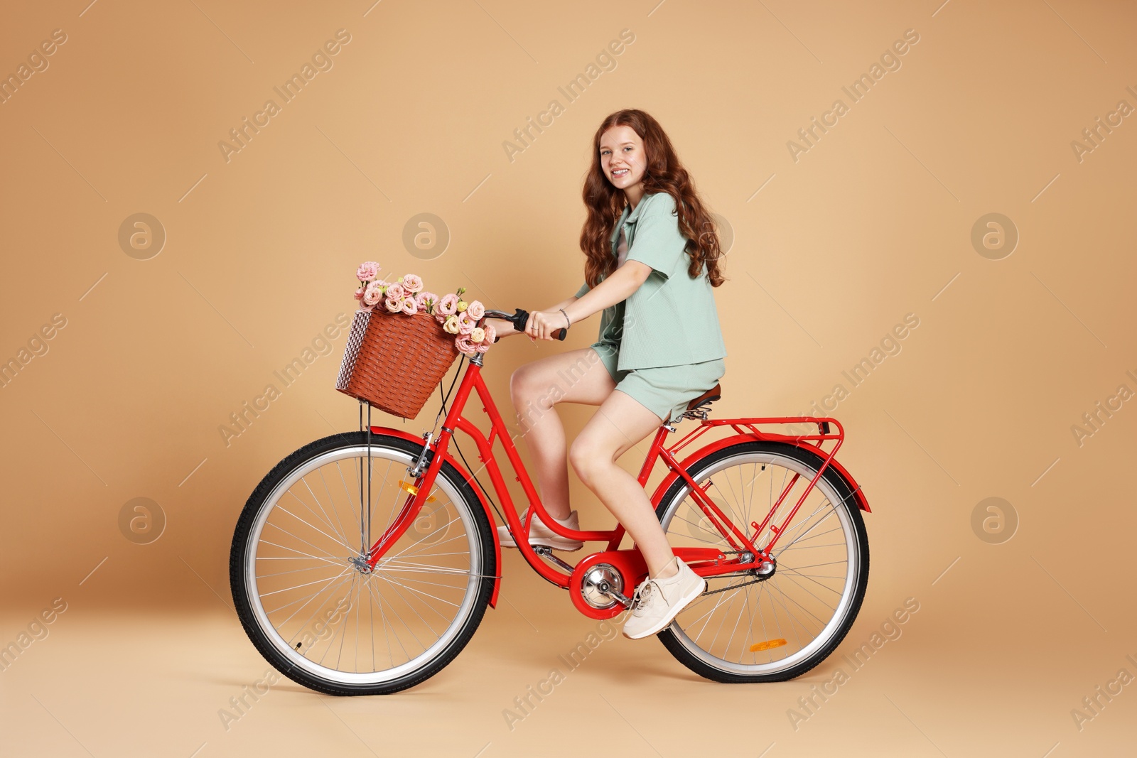 Smiling teenage girl riding bicycle with basket of flowers against beige background Photo of Smiling teenage girl riding bicycle with basket of flowers against beige background