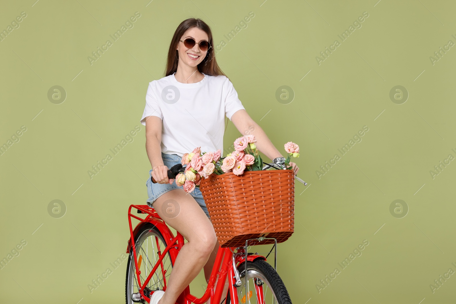 Photo of Smiling woman in sunglasses riding bicycle with basket of flowers on green background