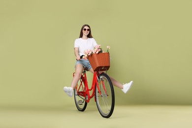 Photo of Smiling woman in sunglasses riding bicycle with basket of flowers on green background