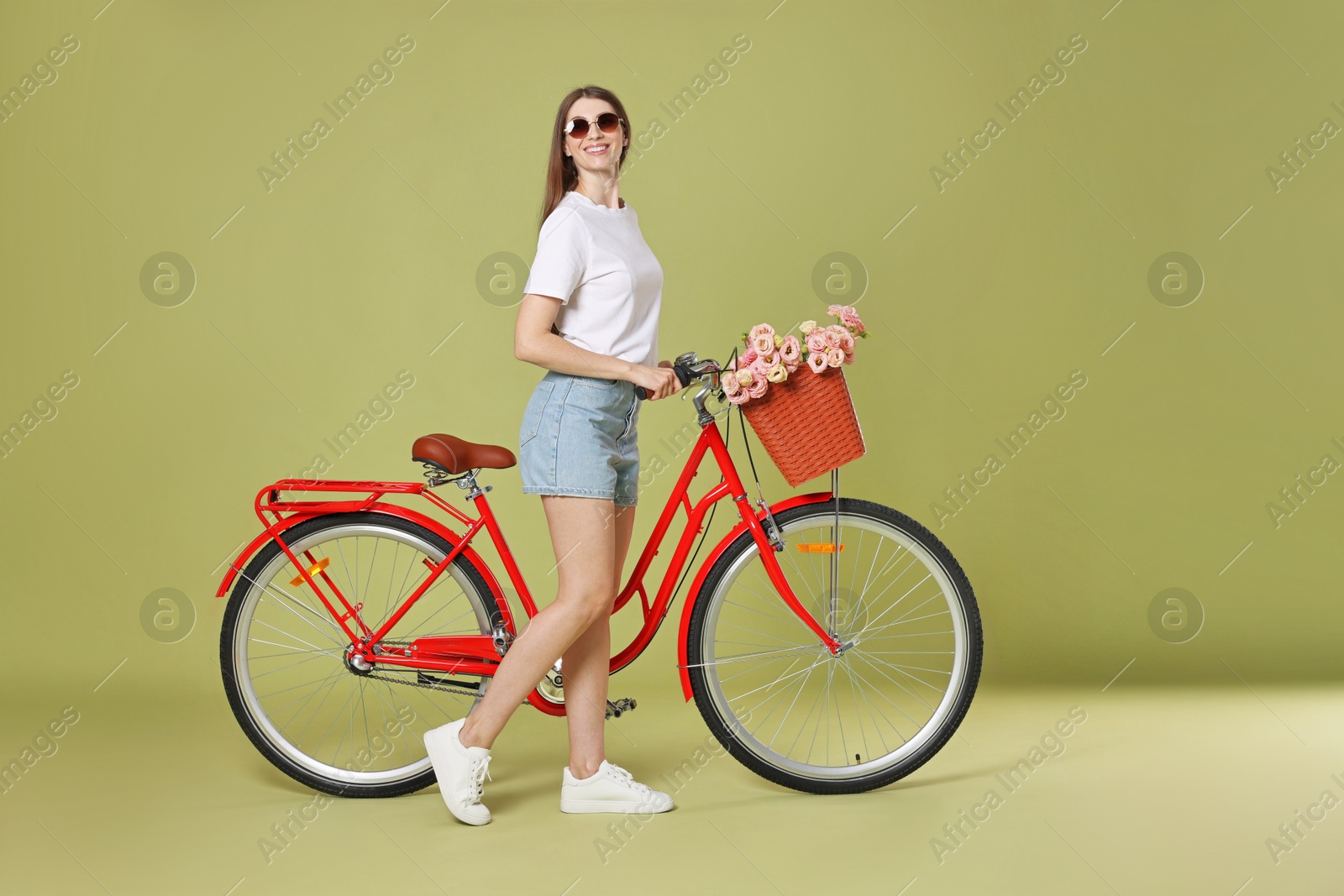 Photo of Smiling woman in sunglasses and bicycle with basket of flowers on green background