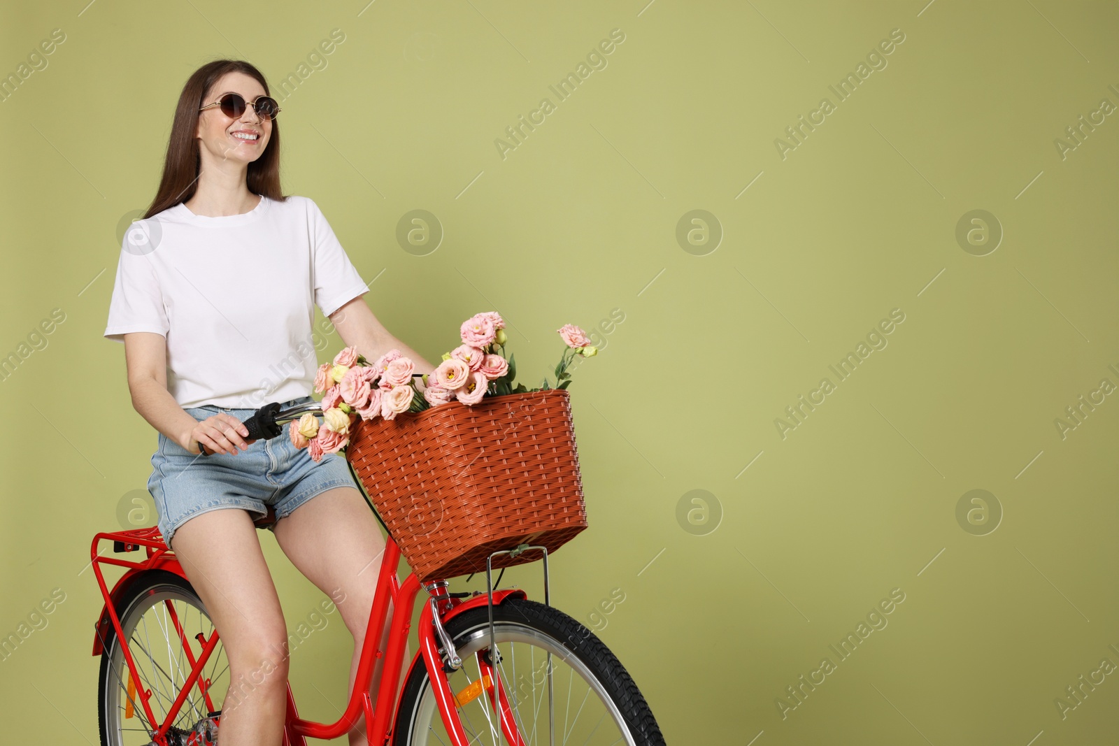 Photo of Smiling woman in sunglasses riding bicycle with basket of flowers on green background. Space for text