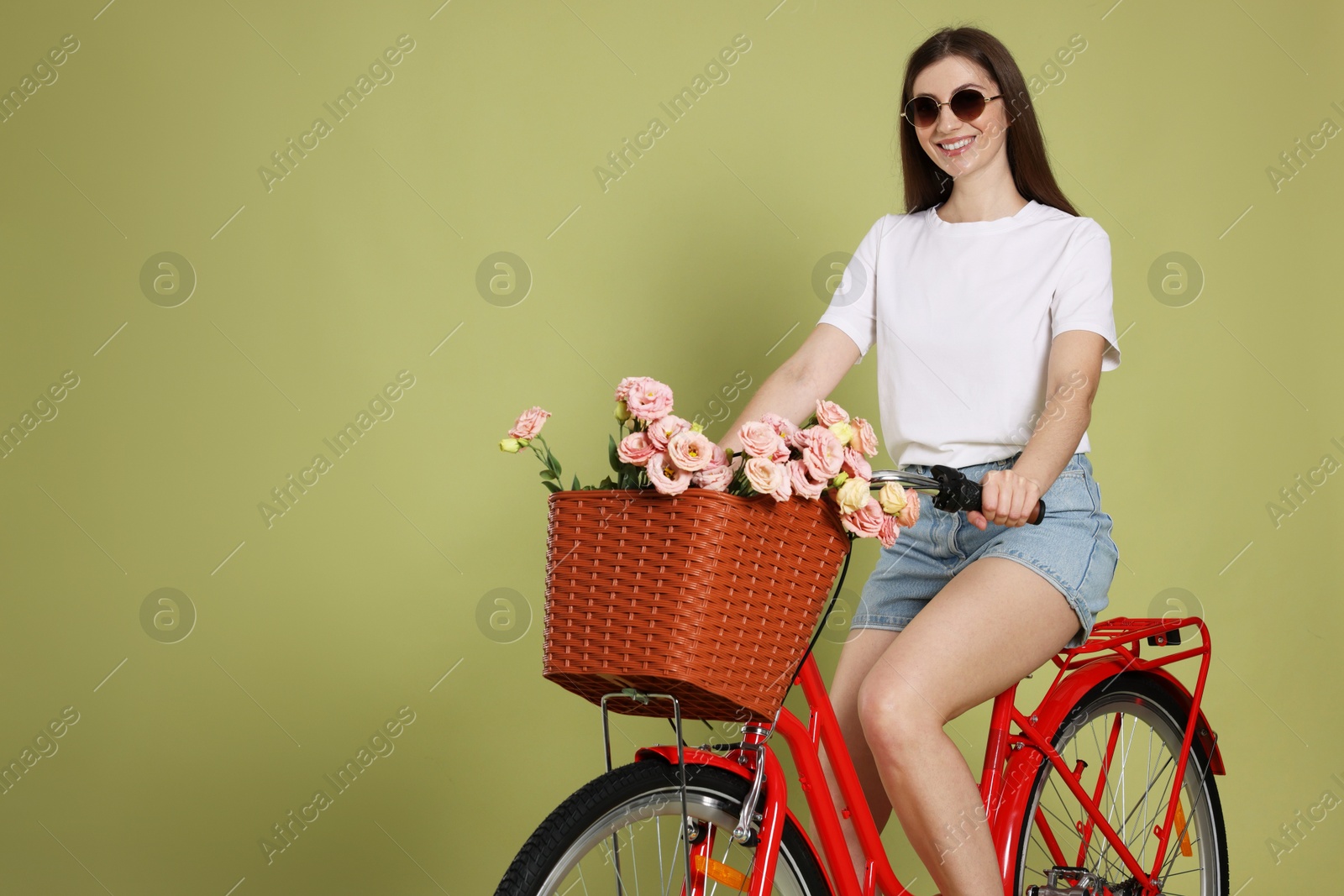 Smiling woman in sunglasses riding bicycle with basket of flowers on green background. Space for text Photo of Smiling woman in sunglasses riding bicycle with basket of flowers on green background. Space for text