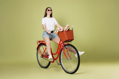 Photo of Smiling woman in sunglasses riding bicycle with basket of flowers on green background