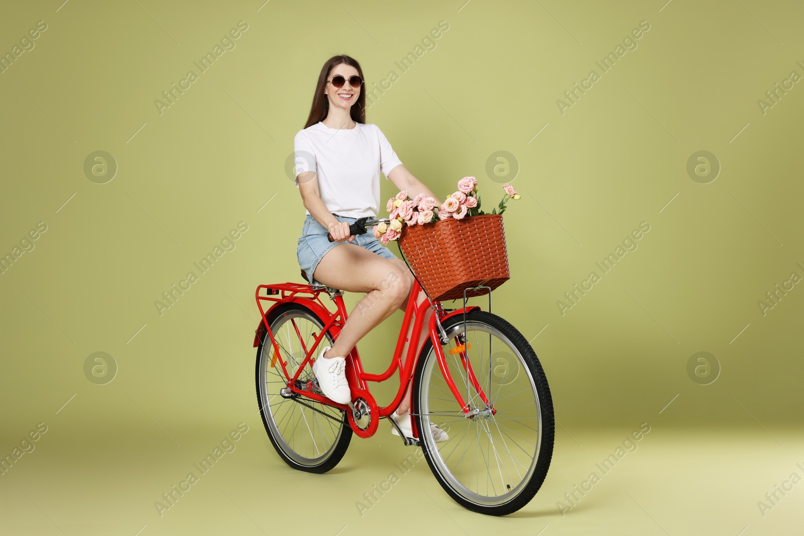 Photo of Smiling woman in sunglasses riding bicycle with basket of flowers on green background