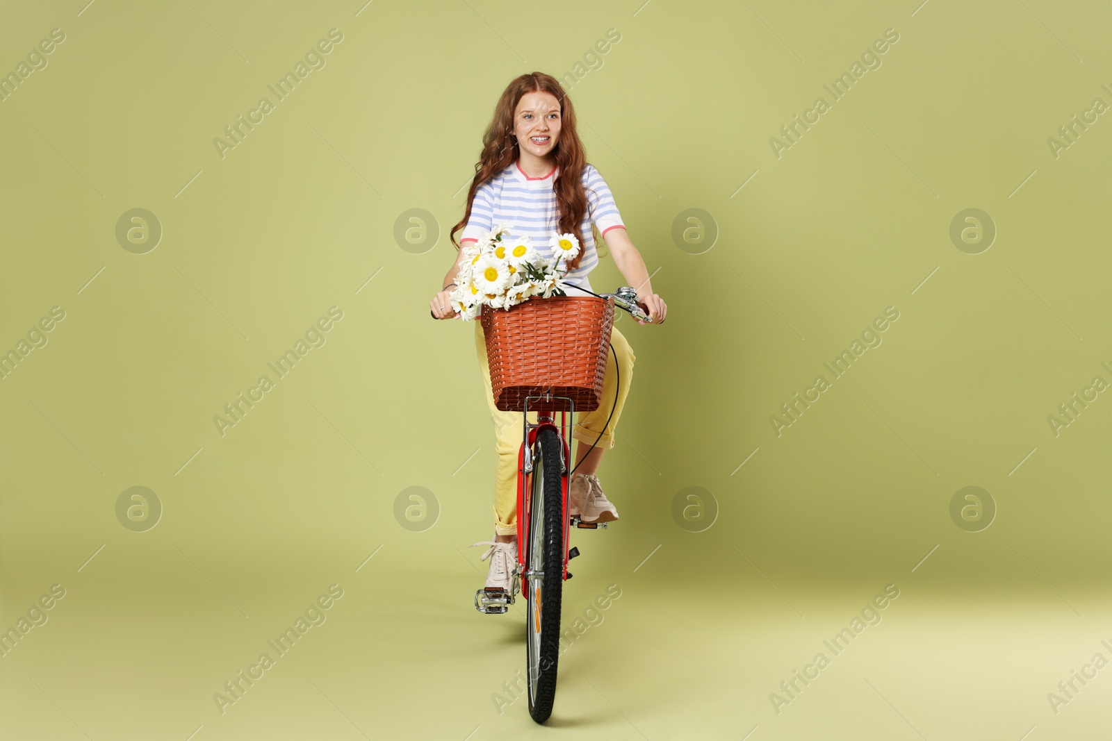 Smiling teenage girl riding bicycle with basket of flowers against green background Photo of Smiling teenage girl riding bicycle with basket of flowers against green background