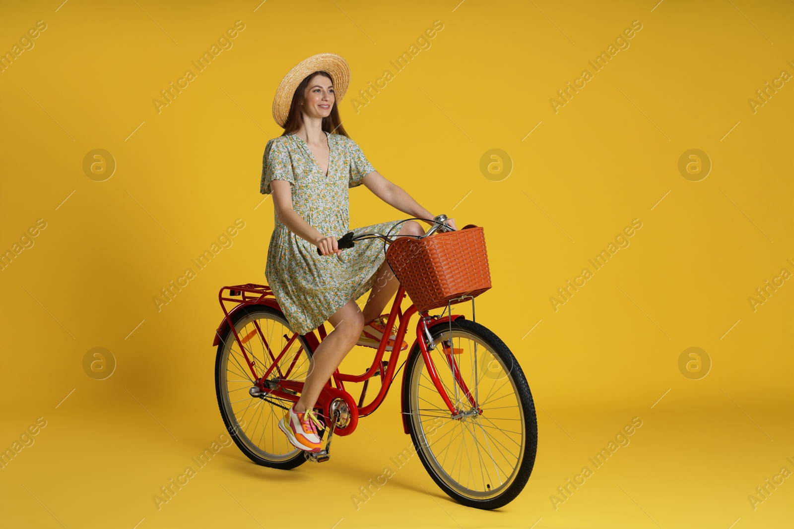 Smiling woman in hat riding bicycle with basket against yellow background Photo of Smiling woman in hat riding bicycle with basket against yellow background